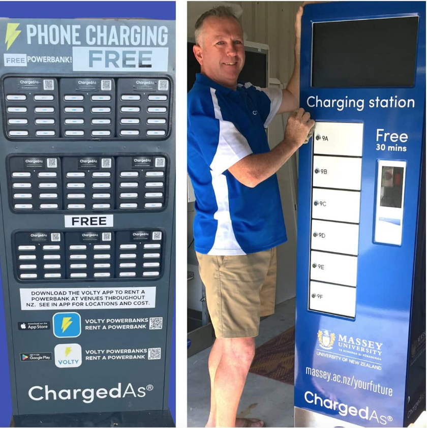 Left, a black cabinet containing three rows of charging stations containing white power banks which has a lightning symbol next to the words "FREE PHONE CHARGING" at the top and the word "FREE" in the middle. Some more words that are too small to read are displayed below the bottom row. The yellow lightning bolt and sky-blue background that represents the Volty app logo is displayed below that; right, a smiling man with greying hair and blue eyes wearing a blue top with a white stripe down the side and beige shorts rests his right fist on a mostly blue phone charging station that says in white "charging station" at the top below an LCD screen and "free 30 mins" below that. The six lockers below the word "charging station" are white. Massey University's logo can be seen below that and below that the word "ChargedAs" in white.