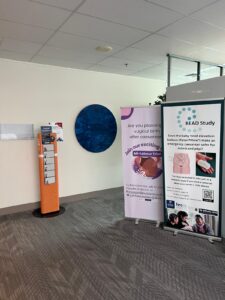 From left; an orange phone charging station with six white lockers on a grey-carpeted floor in Auckland City Hospital, with a blue sign with white writing on it saying "phone charging"; a sign about vaginal births showing a hand on a pregnant woman's stomach; a sign about a BEAD Study