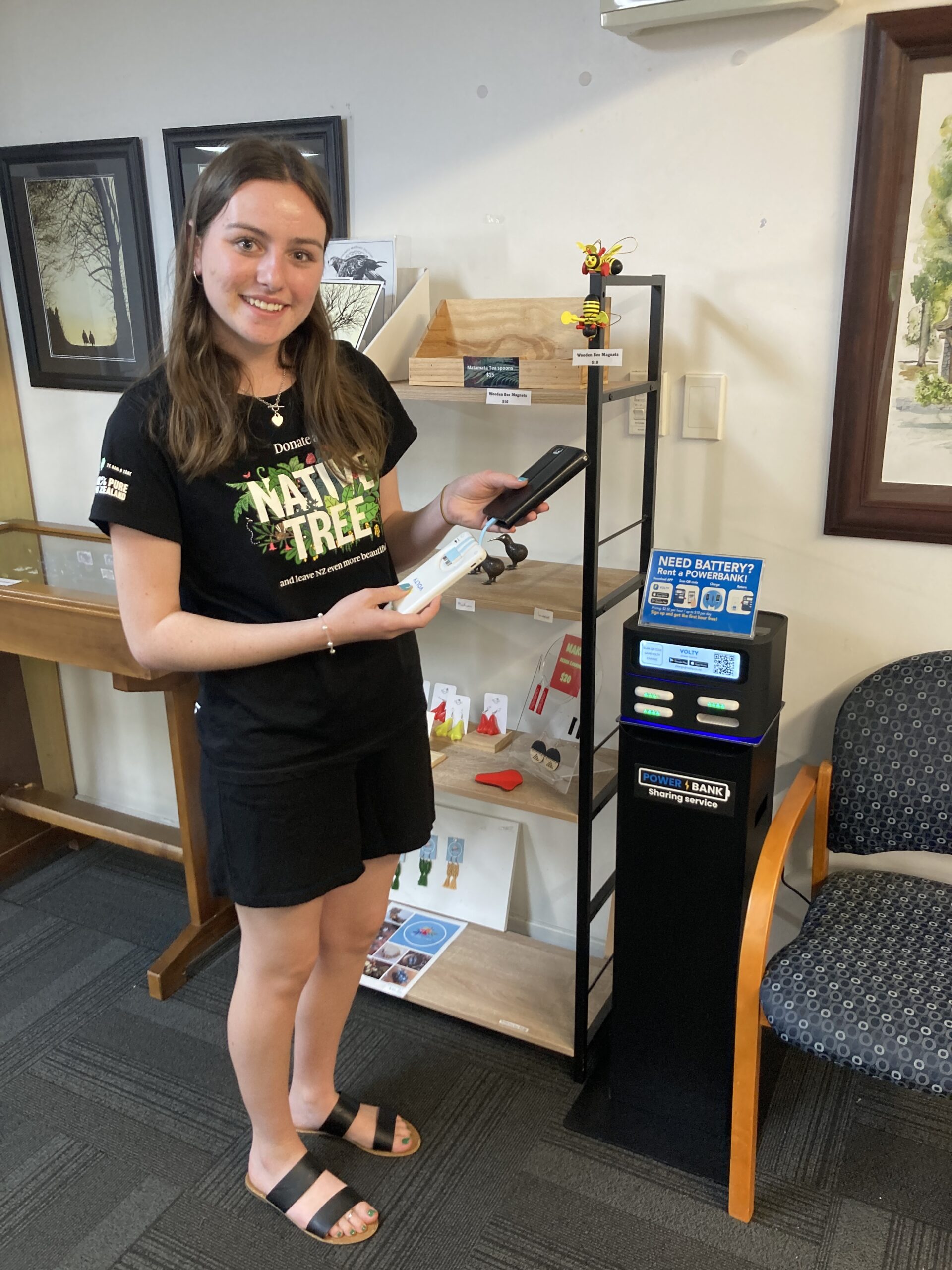 A young woman with long, dark hair below her shoulders and dressed in a black top and skirt, with the words "NATIVE TREE" emblazoned across her chest in yellow with a green background, smiles as she holds her black phone, which is being charged by a white Volty Powerbank with blue cables. She is standing beside a black Volty Powerbank station with three white power banks in the four slots.