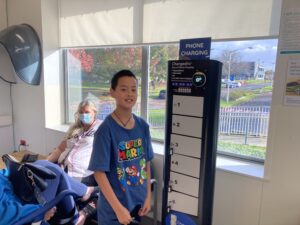 A boy of Southeast Asian origin, aged about 10, dressed in a blue t-shirt with Super Mario on it, grins as he stares into the camera and holds a blue ANZ bank card, beside a six white lockers with keys in a black ChargedAs phone charging station with a blue sign featuring words "PHONE CHARGING" in white. A grey-haired seated white woman, wearing a blue face mask and pink blouse looks into the camera.