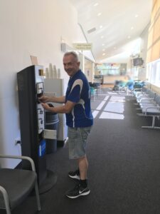 A man dressed in a blue and white t-shirt with the ChargedAs logo, who has short, greying hair, grins as he inserts his black phone to the second-top white locker in a six-locker black charging station of an emergency department with grey chairs behind it. Blurred faces can be seen in the distance.