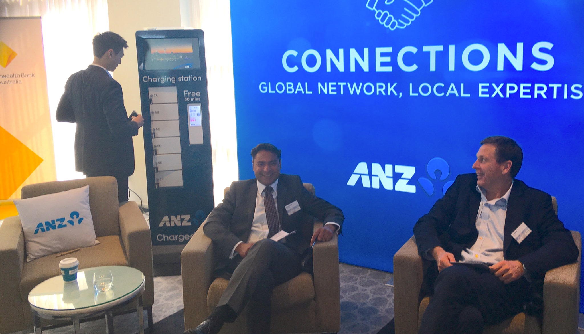Left, a man in a navy suit uses one of six white lockers on a phone charging station that's wrapped in ANZ's blue branding as a video plays on the screen; right, two men in black jackets, white shirts and blue ties smile while sitting in lounge-style chairs in front of an ANZ sign that also features the word "connections" in white writing against a blue background.