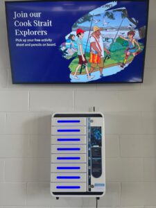 A white, six-locker phone charging station with blue panels stands below a sign featuring a cartoon of travellers with the words "Join Our Cook Strait Explorers" above it.
